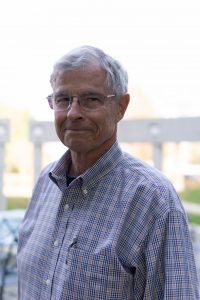 A headshot of Dr. Tom Bacon who is standing outside in front of a grey fence. Tom is wearing a pair of frameless rectangular glasses and a patterned pink and purple collard shirt.