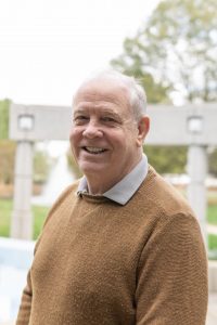 A headshot of Tom Ricketts who is standing outside in front of a grey fence and trees in the far background. Tom is wearing a light brown sweater with a grey collard shirt underneath. 