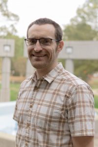 A headshot of Evan Galloway who is standing outside in front of a grey fence and trees in the far background. Evan is wearing a short sleeved plaid tan and brown collard shirt. Evan has short brown hair and is wearing thin, dark-framed square glasses.