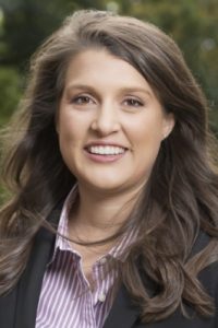 A headshot of Brianna Lombardi. Brianna has long brown wavy hair in this image and wearing a pink white and striped collard shirt under a black blazer.