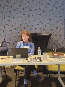 An image of Dr. Erin Fraher, sitting behind a desk presenting. Dr. Fraher has red curly hair and is wearing a blue dress shirt. She is sitting in a chair with a white table in front of her. On the table are stacks of papers, water bottles and a name tag with her name written on it. She is wearing black pants, polka dot socks (grey socks with pink, blue, yellow and black dots) and black dress shoes.