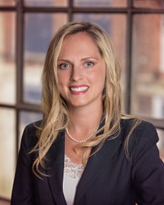 A headshot of Catherine Moore. Catherine has mid-length blonde hair and is wearing a black blazer and white dress shirt. She is smiling directly at the camera in front of a large window. 