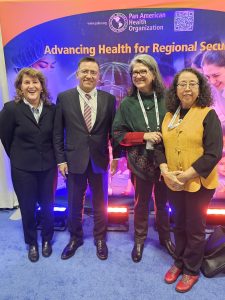 Four people standing and posed in front of a Pan American Health Organization Banner. 