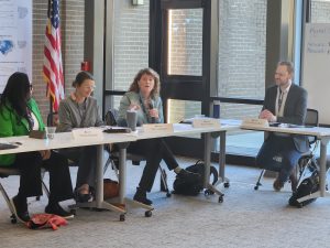 Erin Fraher presenting with a microphone, seated behind a table next to 3 other panel presenters. 
